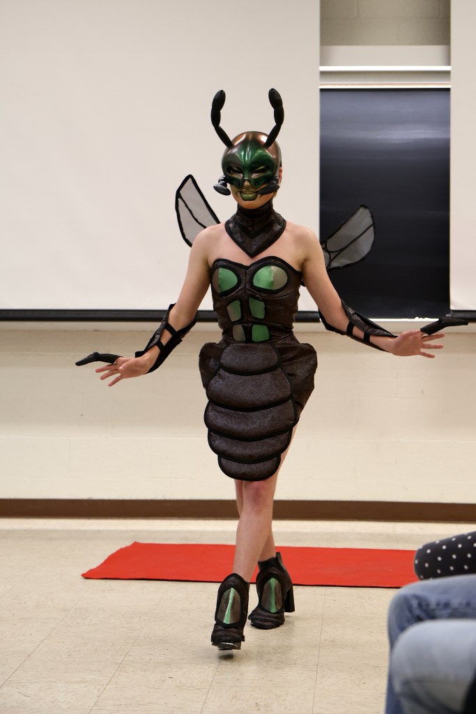 Eldra Arts struts gracefully while wearing a green beetle-inspired costume. The skirt is brown faux leather sewn into a segmented structure reminiscent of an insect abdomen. The corset, heels, and mask are all shiny metallic green. The mask has antennae and palps and covers the top half of Eldra's face. On the bottom half of her face is a thick green lipstick. Two gray wings stick out behind her back. 