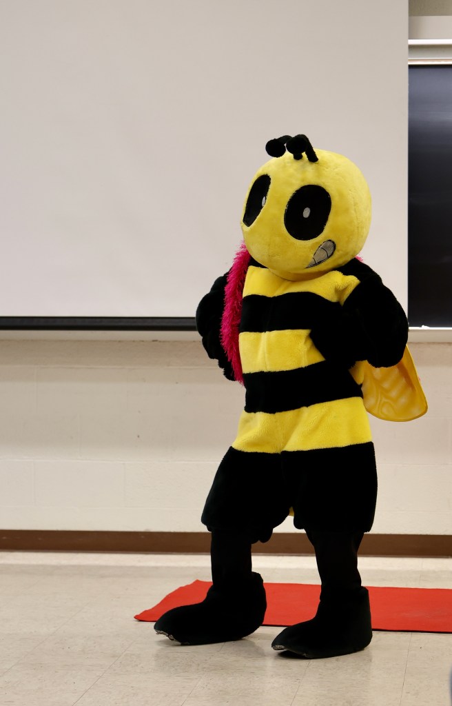 A yellow and black mascot costume of a grinning bee stands jauntily with her hands on her hips. A fluffy pink boa is draped over her shoulder. 